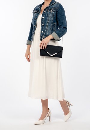 Pochette noire avec un accent métallique en forme de V, associée à une robe midi blanche et une veste en jean. Des chaussures blanches à talons complètent le look.