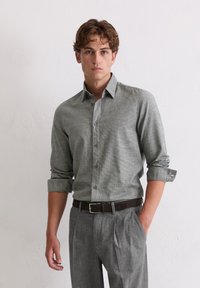 Graues gemustertes Hemd mit langen Ärmeln, die an den Ärmeln hochgeschlagen sind, kombiniert mit grauen Hosen und einem braunen Gürtel. Glatte Stofftextur.