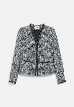 Femme portant un tailleur jupe gris texturé avec une bordure noire, un haut noir, tenant une pochette noire et portant des talons hauts noirs.