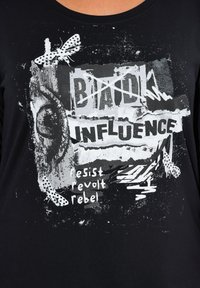 T-shirt noir en coton avec un design graphique en blanc et gris portant le texte : "MAUVAISE INFLUENCE," entouré de motifs abstraits et d'une illustration de papillon.