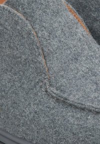 Szare pantofle wsuwane o zaokrąglonym kształcie, z przeszyciami, brązowymi skórzanymi akcentami i teksturowaną powierzchnią. Widziana gumowa podeszwa na spodzie.