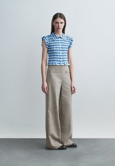 Μια γυναίκα στέκεται μπροστά σε απλό φόντο, φορόντας μπλε και λευκό καρό αμάνικο μπλουζάκι με βολάν και μπεζ παντελόνι με φαρδιές μπατζάκια, συνδυασμένο με μαύρα παπούτσια.