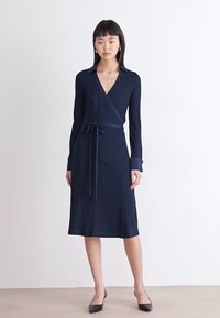 Navy blue ribbed dress with a v-neck, long sleeves, wrap design, and a tie belt at the waist. Features a knee-length hem and structured collar.