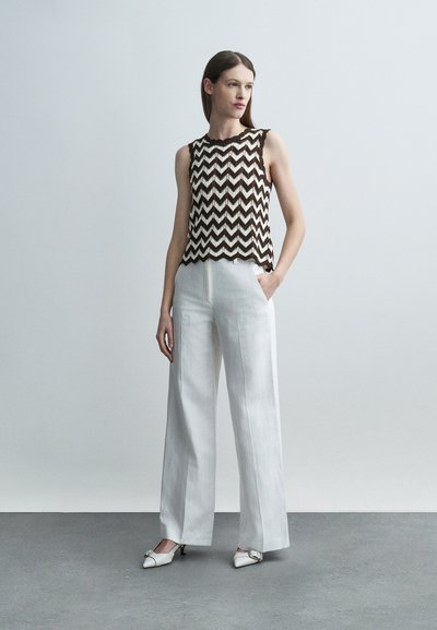 Γυναίκα με αμάνικη μαύρο-λευκή μπλούζα με σχέδιο ζιγκ-ζαγκ, λευκό παντελόνι φαρδύ και άσπρα παπούτσια με χαμηλό τακούνι, στέκεται μπροστά σε απλό γκρι φόντο.