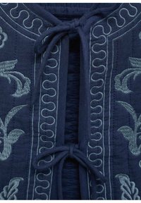 Navy tkanina oblačilo z zapletenim svetlo modrim vezenjem. Oblikovanje vključuje cvetlične vzorce in dekorativno zavezovanje pri ovratniku.