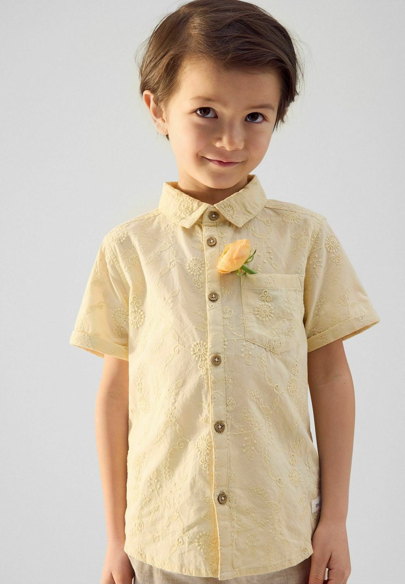 Korte mouwen geel shirt met bloemenborduursel, knoopsluiting aan de voorkant en een borstzak. Heeft een bloemenaccent vastgepind aan de kraag.