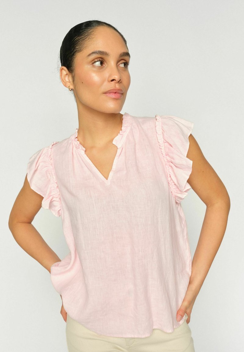 Sötét hajú nő alacsony konttyal, világos rózsaszín fodros ujjatlan blúzt és bézs nadrágot visel, kissé oldalra néz.