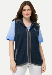 Námořnický pletený vest s kapucí, šedými akcenty, předním zipem a kapsami. Nosený přes světle modrou košili s kulatým výstřihem.