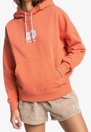 Personne portant un sweat à capuche corail avec un imprimé floral et un short en velours côtelé beige, main dans la poche du sweat, debout devant un fond blanc.