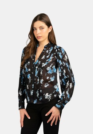 Blusa nera a fiori con pattern blu e bianchi, tessuto semi-trasparente, maniche lunghe, dettagli plissettati e chiusura con bottoni. Abbinata a pantaloni neri.