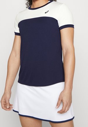 Person wearing a navy and white short-sleeve sports top with logo, paired with a white skirt featuring navy trim at the hem.