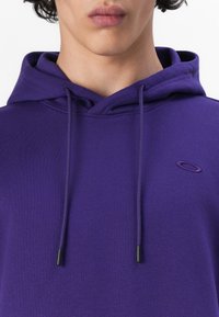 Sudadera morada con capucha ajustable, de tejido suave, y un pequeño logo bordado en el pecho, con extremos de los cordones en negro.