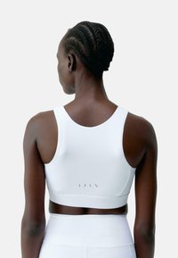Bijela sportska bralette od glatkog, rastezljivog materijala, s niskim dizajnom na leđima i minimalističkim logotipom na elastičnom dijelu.