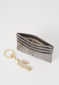 Portafoglio grigio lucido con motivo geometrico in rilievo; dotato di più scomparti e una tasca interna. Include un portachiavi dorato con un design a forma di mano decorato con strass.