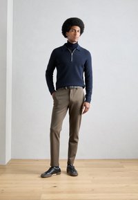 Marineblå strikkegenser med glidelås krage, kombinert med beige bukser og svarte joggesko med hvite detaljer. Modellen står på et tregulv.