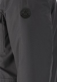 Svart tygjacka med en mjuk struktur har en cirkulär svart emblem på ärmen med en bergdesign och texten "WHISTLER".