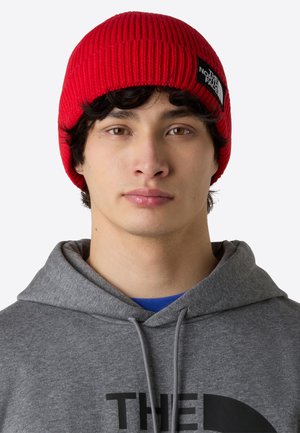 Gorro de punto acanalado rojo con una placa de logotipo negra; llevado con una sudadera gris. El gorro se ajusta perfectamente, mostrando una superficie texturizada.