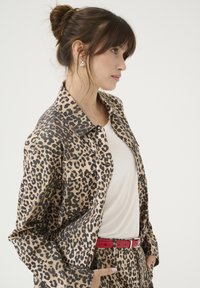 Giacca a camicia con stampa leopardata in beige e nero, con chiusura a bottoni, due tasche frontali, indossata sopra una maglietta bianca, completata da una cintura rossa.