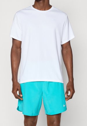 Mężczyzna ubrany w gładką białą koszulkę z krótkim rękawem i turkusowe sportowe szorty z białymi logo Nike na piersi i na nogawce.