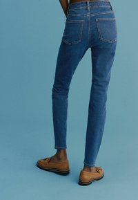 Sininen farkut, joissa on slim fit, korkea vyötärö ja takataskut. Yhdistetty ruskeisiin loafereihin, vaaleansinistä taustaa vasten.