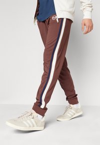 Bruna joggerbyxor med marinblå och vita sidostripes, matchande ribbad mudd, som bärs med beige sneakers. Tyget verkar mjukt och elastiskt.