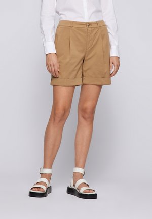 Kvinde iført beige shorts med opbuk, hvid langærmet skjorte og hvide sandaler med spænde, stående foran en ensfarvet grå baggrund.