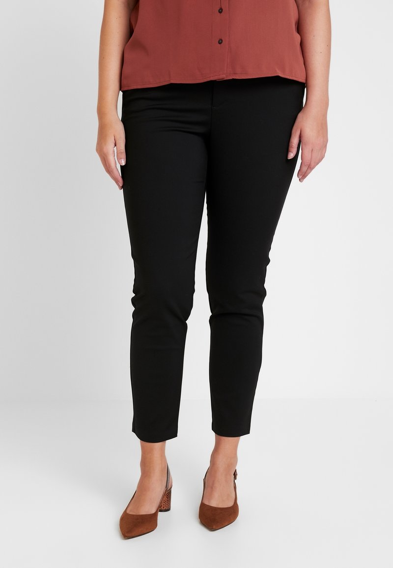 Zwarte, slim-fit broek met een gladde textuur, gecombineerd met een roestkleurige blouse met knopen aan de voorkant en korte mouwen. Bruine schoenen met blokhak.