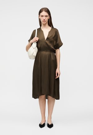 Naine, kes kannab pruuni põlvikuni kleiti, musti teravate ninadega kingi ning hoiab valget õlakotti, taustaks hele ühtlane sein.