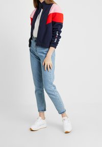 Námořnická bomber bunda s červenými, růžovými a bílými geometrickými barevnými bloky. Spárovaná s světle modr ými džíny a bílými teniskami, stojící na bílém pozadí.