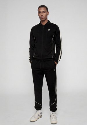 Homme debout, les mains dans les poches, portant un survêtement noir avec des bandes blanches sur les côtés et des baskets blanches sur un fond gris uni.