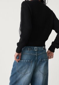 Zwarte geribbelde gebreide trui met omgeslagen mouwen, gecombineerd met lichtblauwe denim jeans met dubbele achterzakken en een zichtbaar riemlusdetail.