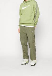 Zelená mikina Nike s velkým bílým logem, doplněná o zelené tepláky ve stejném odstínu. Oba kousky mají volný střih a detaily na šňůrkách. Bílé akcenty.