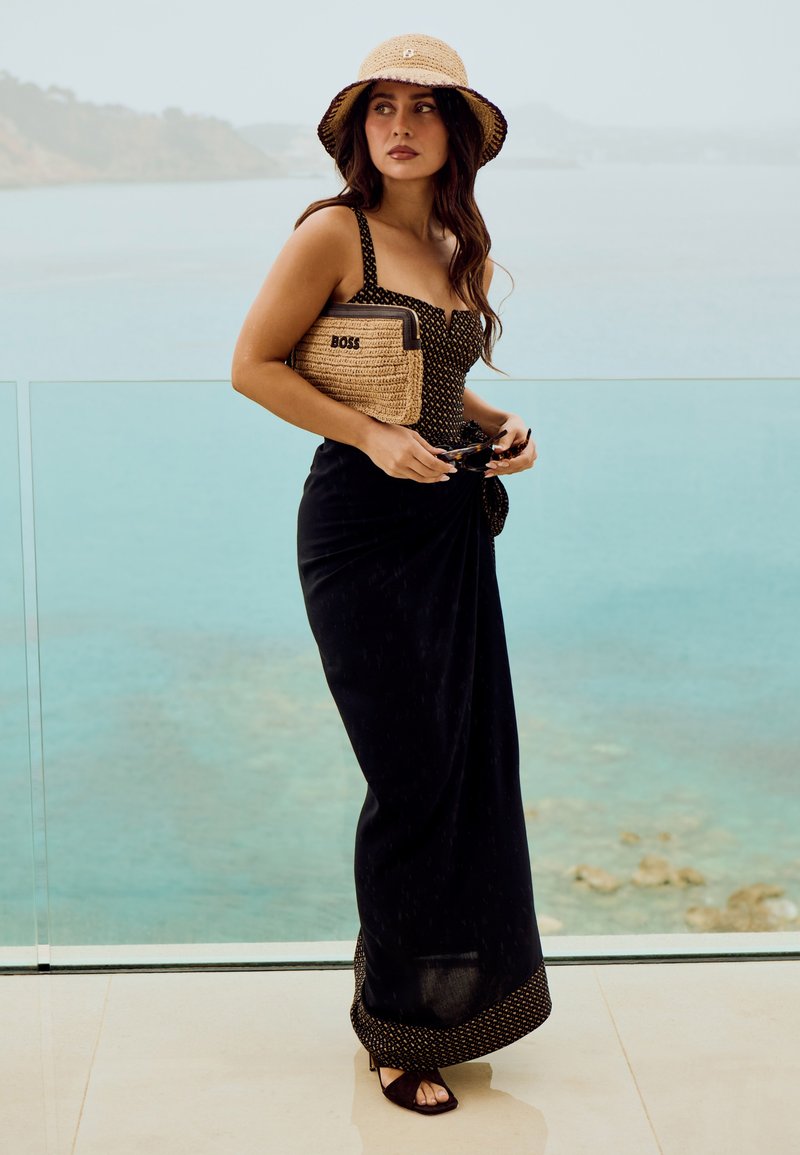 Femme en robe noire et chapeau de paille tenant une pochette tissée et des lunettes de soleil, debout près d'une rambarde en verre avec vue sur la mer.