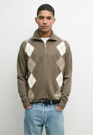 Junger Mann trägt einen braunen Pullover mit Rautenmuster, Halbreißverschluss und hellblauen Jeans, steht mit den Händen in den Taschen.