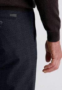 De hand van een man naast de zak van een donkergrijze ruitjesbroek met een klein metalen label, terwijl hij een donkerbruine lange mouwen shirt draagt.