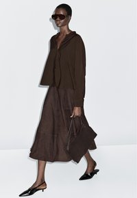 Brązowa warstwowa bluzka i spódnica wykonane z miękkiego materiału, zestawione z czarnymi sandałami na obcasie z odkrytą piętą i spiczastym noskiem oraz dużymi ciemnymi okularami przeciwsłonecznymi. Nosi ciemną torebkę.