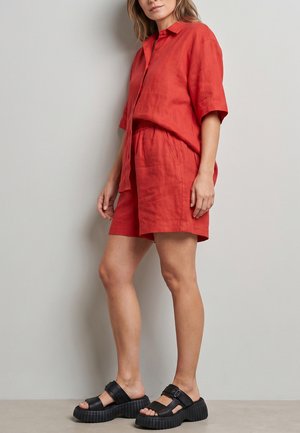 Vrouw draagt een losse rode blouse met knopen en bijpassende shorts met zwarte platformsandalen, staande tegen een effen lichtgrijze muur.