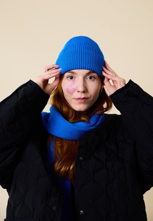 Blue knitted beanie and matching scarf, textured fabric; black quilted jacket, holding beanie. Neutral background emphasizes the colors.
