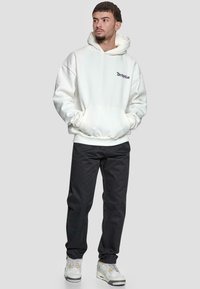 Weißer Kapuzenpullover mit schwarzem Logo, Fronttasche und gerippten Bündchen, kombiniert mit schwarzen Hosen und weißen Sneakers mit grauen Akzenten.