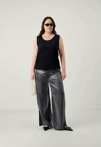 Top nero senza maniche, pantaloni larghi metallici argento, tiene una borsa beige texturizzata, indossa occhiali da sole scuri, stilizzato su uno sfondo neutro.