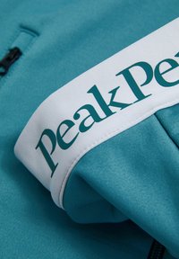 Teal mikina s bielym pruhom s logom "Peak Performance". Tkanina je hladká, s dizajnom na pol zip, a s rebrovanými detailmi.