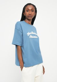 Camiseta de algodão azul com decote redondo e mangas curtas, apresentando o texto branco "New Balance Athletics" na parte da frente.