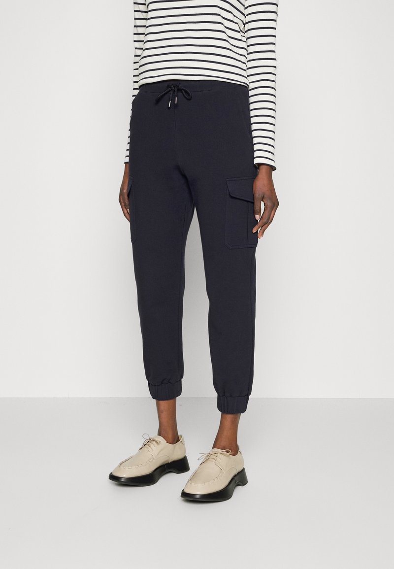 Personne portant un jogger cargo noir avec cordon de serrage aux chevilles, un haut à manches longues rayé blanc et noir, et des chaussures beiges à lacets.