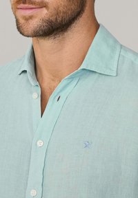 Lichtblauwe linnen blouse met een button-down kraag, voorzien van witte knopen en een klein geborduurd logo op de borst. Zachte textuur.