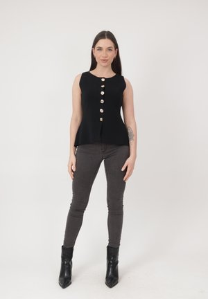 Kvinde med langt mørkt hår iført en ærmeløs sort bluse med knapper, mørkegrå smalle bukser, sorte ankelstøvler og en synlig tatovering på underarmen.