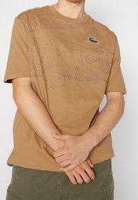 Brun bomulls-T-shirt med en stor grå krokodilgrafik och en liten grön logotyp på bröstet. Korta ärmar, rund halsringning.