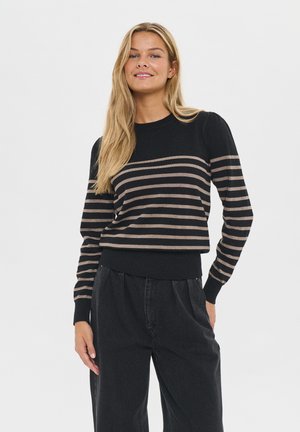 Kvinde med langt blondt hår iført en sort sweater med beige vandrette striber og løse sorte bukser, smilende mod en hvid baggrund.