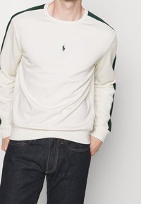 Mikina s kulatým výstřihem v off-white barvě s tmavě zelenými akcenty na ramenou, s malým vyšívaným logem na hrudi. Měkká texturu látky.