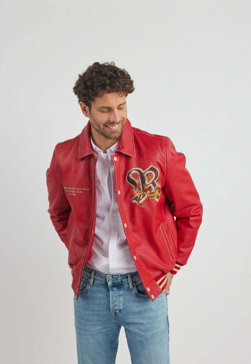 Veste en cuir rouge avec un design brodé "B" proéminent et des accents dorés. Comprend une fermeture éclair et des poignets rayés. Portée par-dessus une chemise blanche.