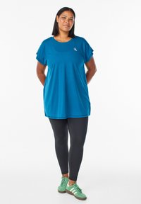 Tirkīza krāsas īsām piedurknēm blūze ar apaļu kakla izgriezumu un nelielu logotipu, apvienota ar melniem legingiem un zaļiem sporta apaviem ar baltām svītrām. Gluda tekstūra.